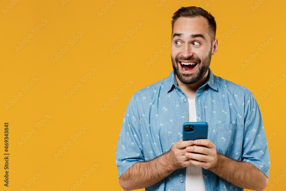 Young smiling happy caucasian man 20s wear blue shirt white t-shirt hold in hand use mobile cell phone chatting browsing look aside on workspace isolated on plain yellow background studio portrait Young smiling happy caucasian man 20s wear blue shirt white t-shirt hold in hand use mobile cell phone chatting browsing look aside on workspace isolated on plain yellow background studio portrait