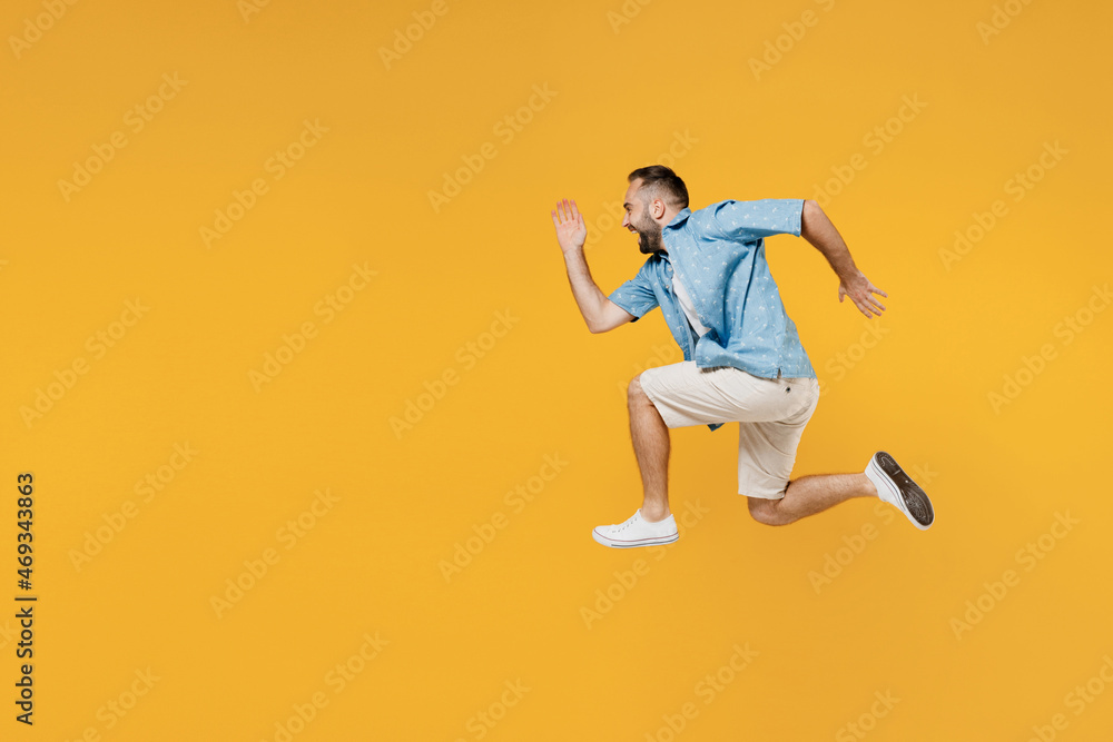 Full body side view young smiling sporty happy caucasian man 20s in blue shirt white t-shirt run fast jump high hurry up isolated on plain yellow background studio portrait. People lifestyle concept.