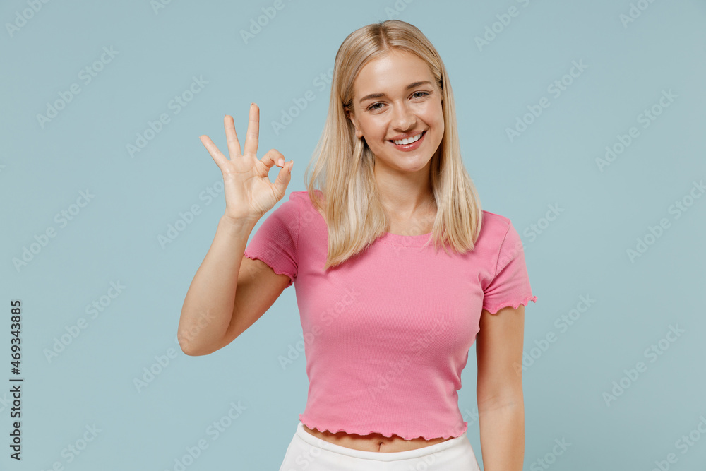 Young smiling happy cheerful blonde woman 20s wearing casual pink t-shirt look camera showing okay ok gesture isolated on plain pastel light blue background studio portrait. People lifestyle concept. Young smiling happy cheerful blonde woman 20s wearing casual pink t-shirt look camera showing okay ok gesture isolated on plain pastel light blue background studio portrait. People lifestyle concept.