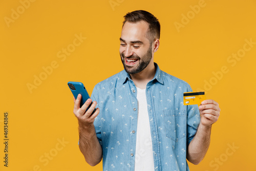 Young happy man 20s wear blue shirt white t-shirt using mobile cell phone hold credit bank card doing online shopping order delivery booking tour isolated on plain yellow background studio portrait