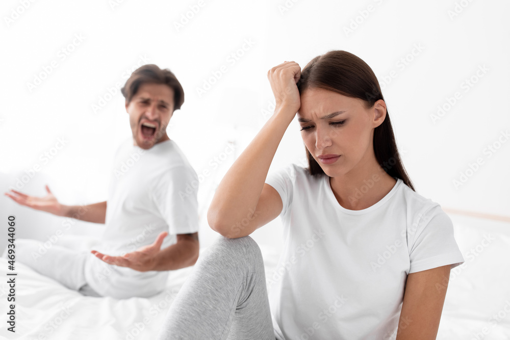 Unhappy upset caucasian millennial wife ignores screaming husband, lady sits in bed in bedroom interior Unhappy upset caucasian millennial wife ignores screaming husband, lady sits in bed in bedroom interior
