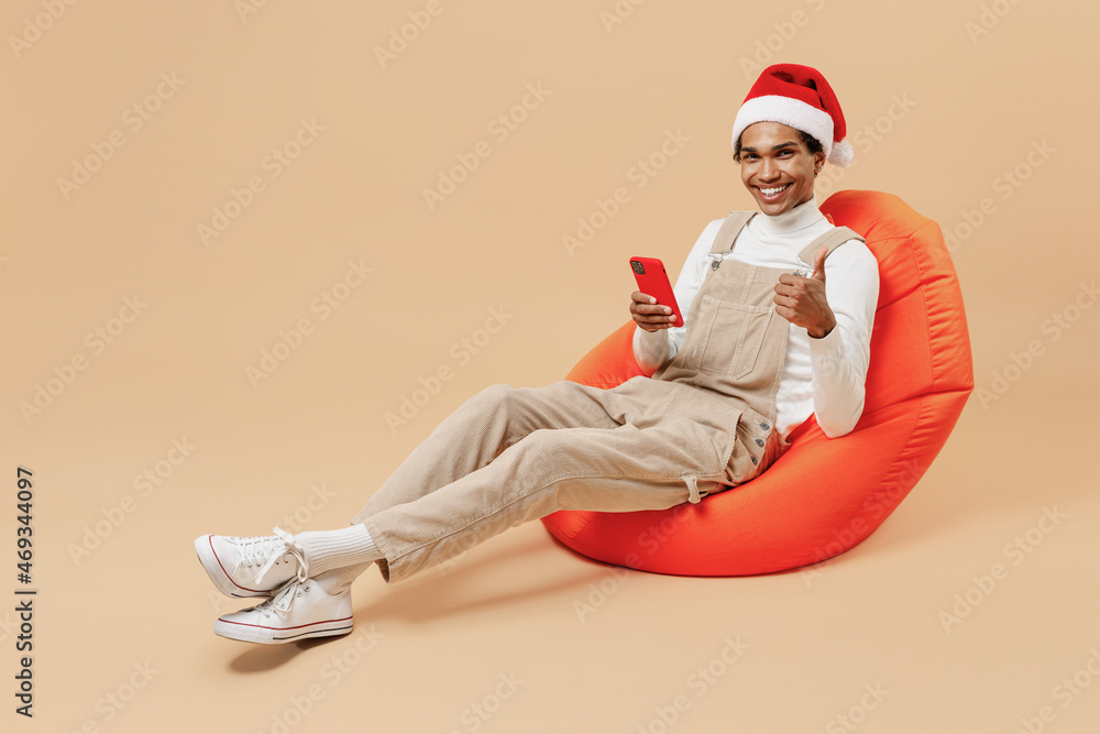 Full size young african man in red Christmas hat sit in bag chair using hold mobile cell phone show thumb up isolated on plain pastel beige background studio. Happy New Year 2022 celebration concept. Full size young african man in red Christmas hat sit in bag chair using hold mobile cell phone show thumb up isolated on plain pastel beige background studio. Happy New Year 2022 celebration concept.