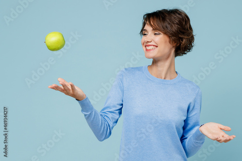 Young smiling satisfied happy vegetarian cool woman 20s in casual sweater look camera toss up green apple isolated on plain pastel light blue background studio portrait. People lifestyle food concept photo