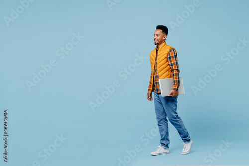 Full size body length fun young black man 20s years old wear yellow waistcoat shirt hold use work on laptop pc computer stepping walking isolated on plain pastel light blue background studio portrait