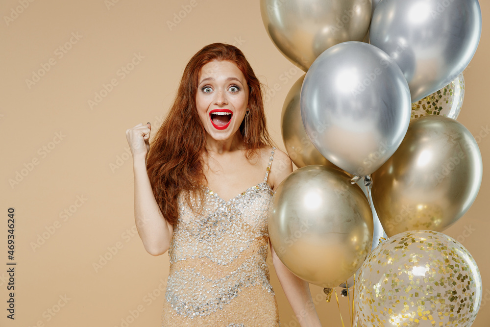 Naklejka premium Young excited redhead woman 20s wearing fancy evening dress hold bunch of air balloons do winner gesture isolated on plain pastel beige background studio portrait. Celebrating birthday party concept.