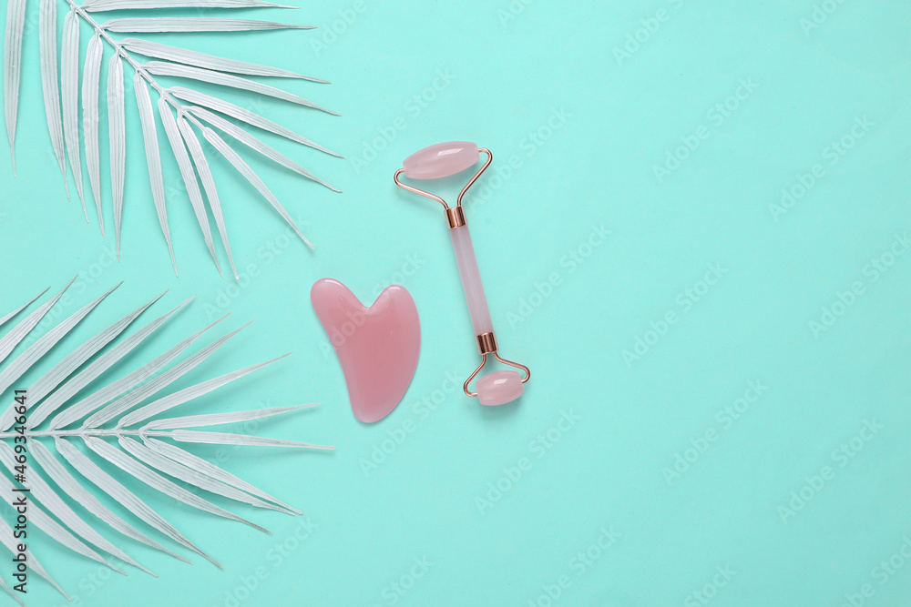 Face roller and gua sha stone massager on blue background with white palm leaves. Blue background. Top view Face roller and gua sha stone massager on blue background with white palm leaves. Blue background. Top view