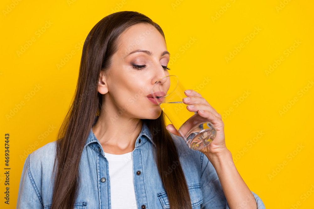 Photo of lovely young lady drink water wear jeans shirt isolated over yellow color background Photo of lovely young lady drink water wear jeans shirt isolated over yellow color background