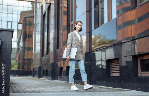Informal, young business woman with a laptop walks through the city on the background of a business building © splitov27