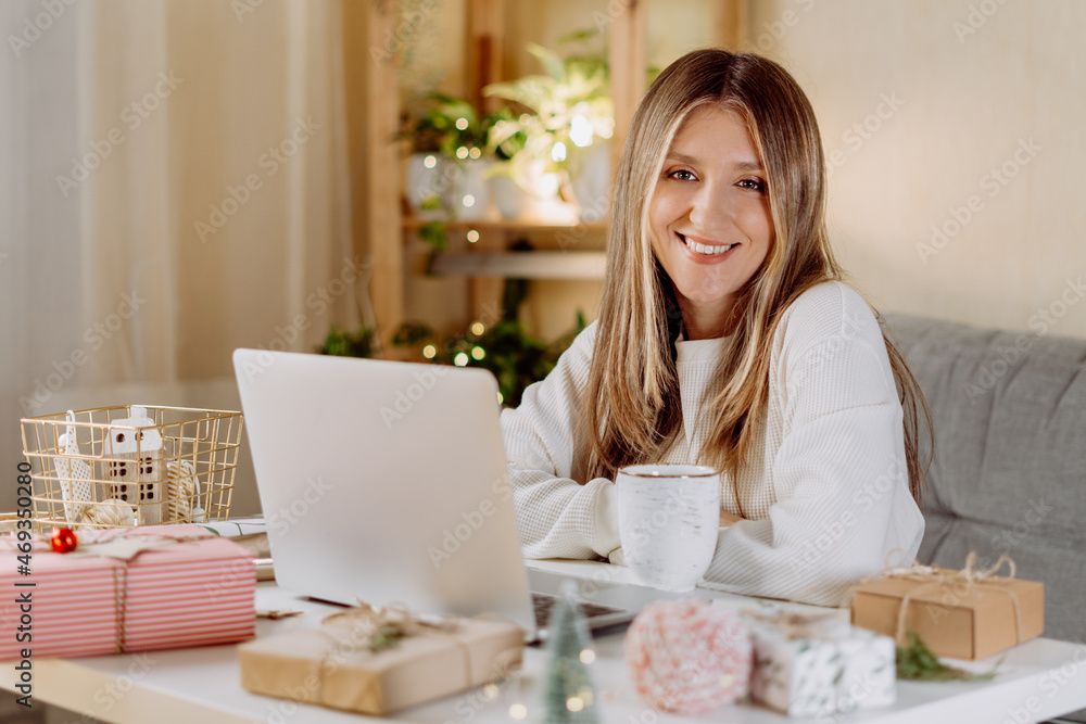 Happy and smiling young woman wrapping and preparing Christmas gifts, typing at laptop and having online video chat. Online shopping at Christmas holidays. Home interior with lights and plants Happy and smiling young woman wrapping and preparing Christmas gifts, typing at laptop and having online video chat. Online shopping at Christmas holidays. Home interior with lights and plants