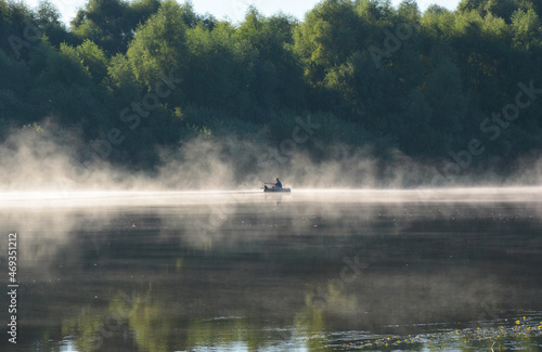 A fisherman on a boat fishing and sailing up the river in the fog. A river landscape with a boat and a green riverbank in summer.