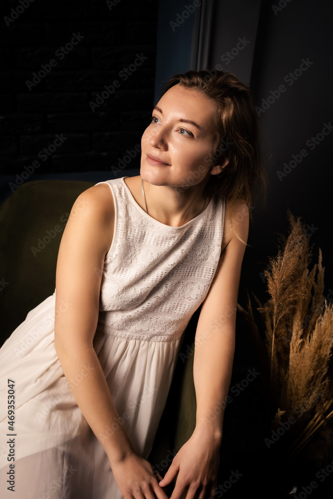 A young woman in a dress with long hair smiles and looks to the side while sitting in a chair in a dark room. A young woman in a dress with long hair smiles and looks to the side while sitting in a chair in a dark room.