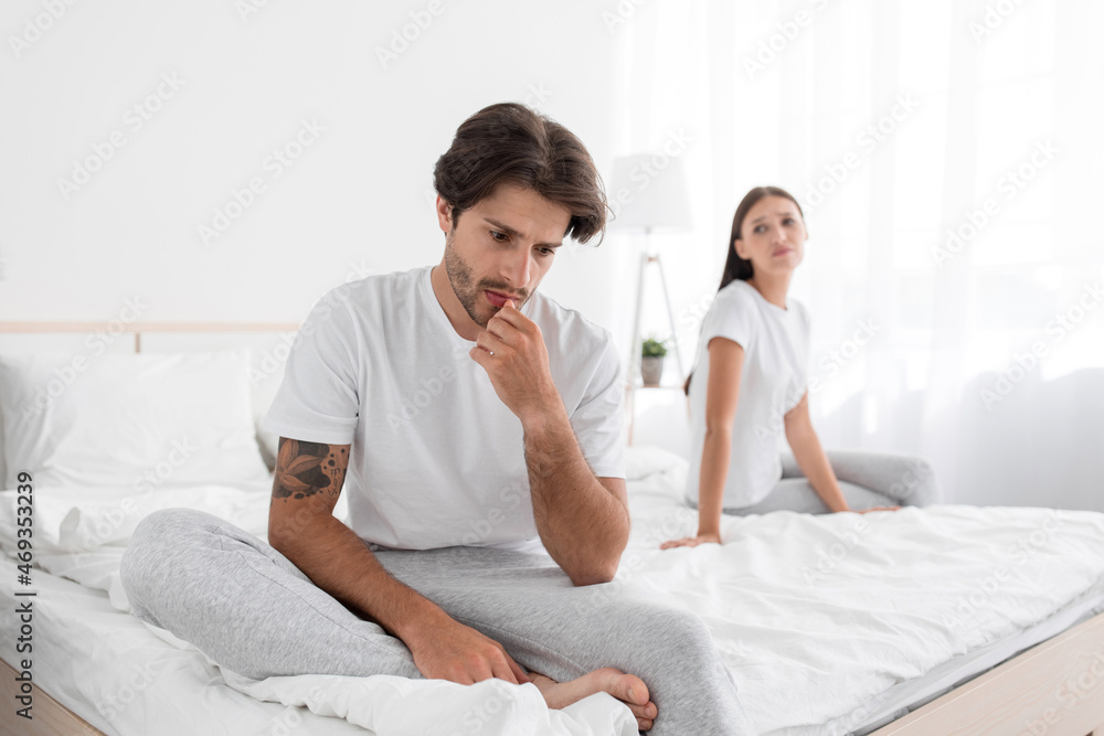 Depressed millennial european husband with stubble sits on bed and sad, wife apologizes in bedroom Depressed millennial european husband with stubble sits on bed and sad, wife apologizes in bedroom