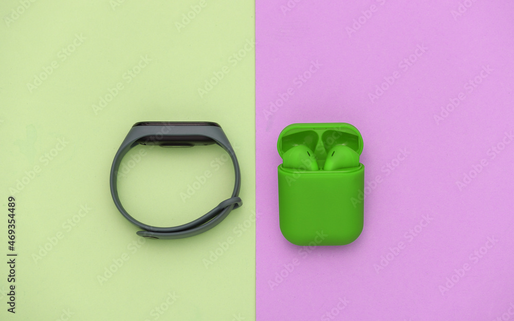 Green wireless headphones with charging case and smart bracelet on pink green pastel background. Top view Green wireless headphones with charging case and smart bracelet on pink green pastel background. Top view