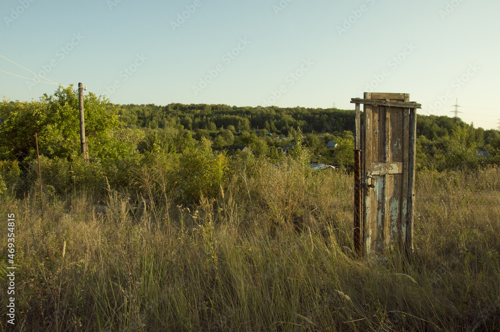 An old wooden door standing in the middle of the road among trees and bushes on a clear cloudless summer evening