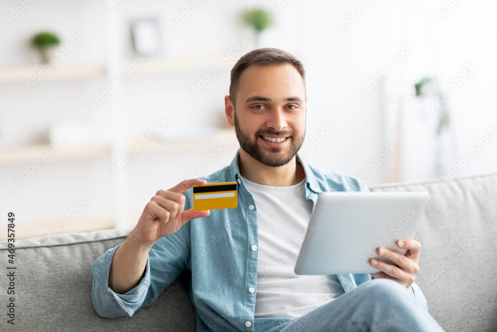 Cheery young guy with credit card and digital tablet sitting on sofa at home, buying goods on web