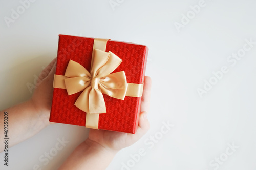 Fototapeta Naklejka Na Ścianę i Meble -  The child holds a red box with a golden bow for a gift on a white background. Decoration for winter holidays, Christmas and New Year.