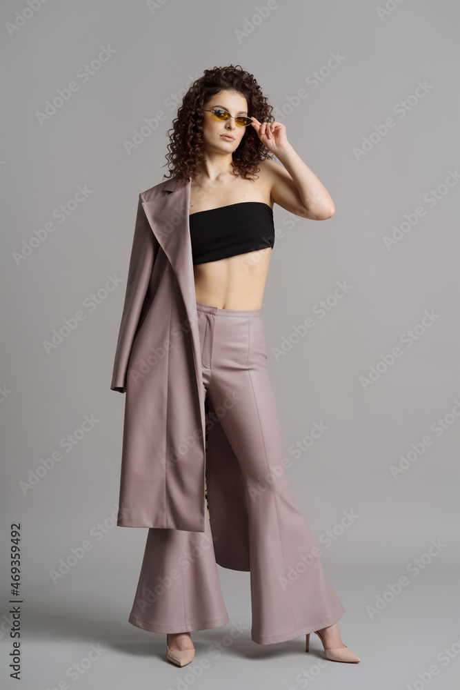 Studio shot of pretty fashion model in pastel purple leather trousers, coat, khaki sunglasses on gray background Studio shot of pretty fashion model in pastel purple leather trousers, coat, khaki sunglasses on gray background