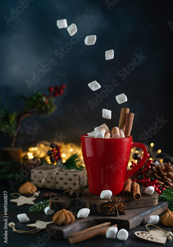 Hot chocolate with marshmallows in a red mug on dark wooden boards. Marshmallows are hanging in the air and scattered on the table. Fir branches  cinnamon tubes  cookies and lights in the background
