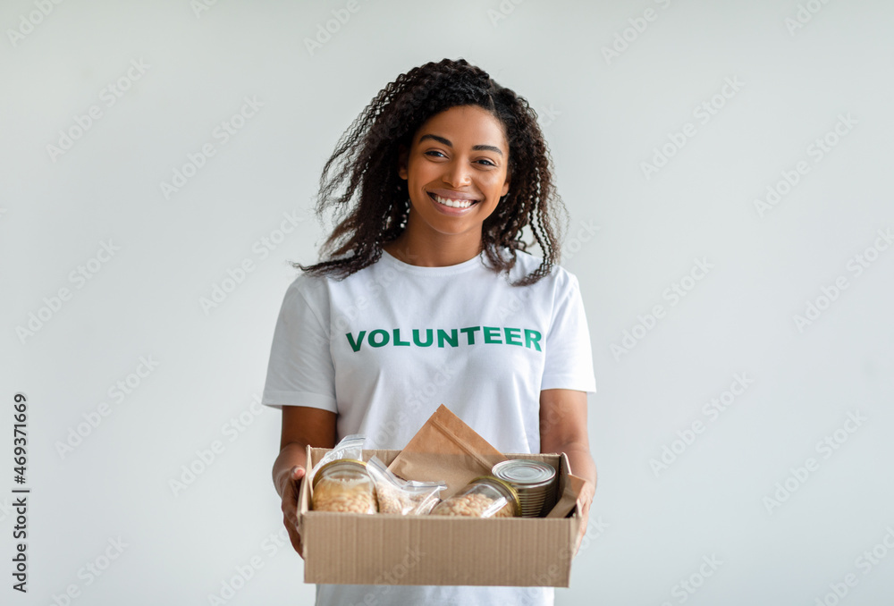 Happy black female volunteer holding donations box with cans and packages, smiling at camera, standing over light wall Happy black female volunteer holding donations box with cans and packages, smiling at camera, standing over light wall