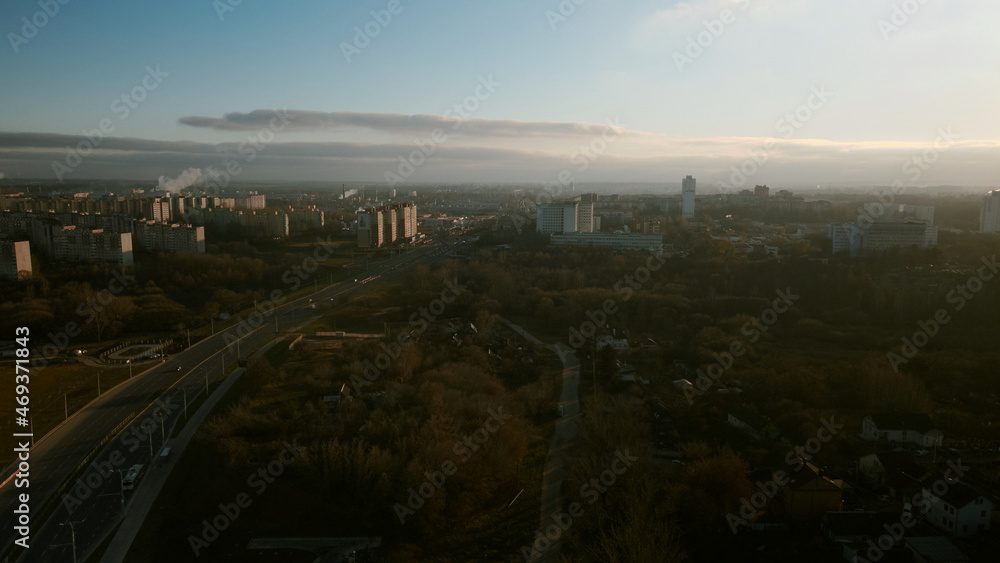 The suburb is backlit by the setting sun. Houses, a park area and a city highway are visible. Aerial photography.