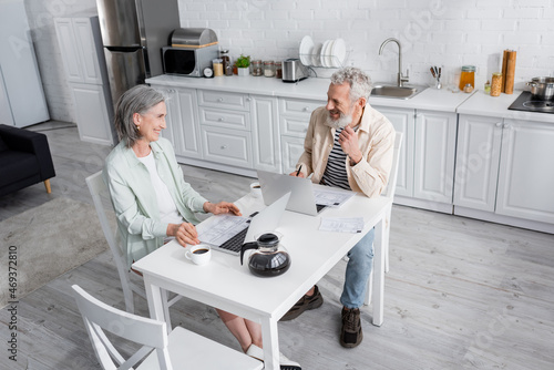Cheerful mature couple looking at each other near bills, coffee and laptops at home.