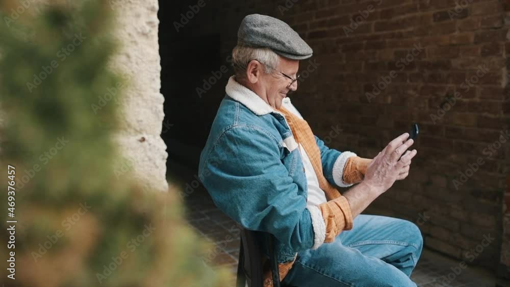 Stylish senior in fashionable clothes and in glasses sits on the chair and uses phone Stylish senior in fashionable clothes and in glasses sits on the chair and uses phone