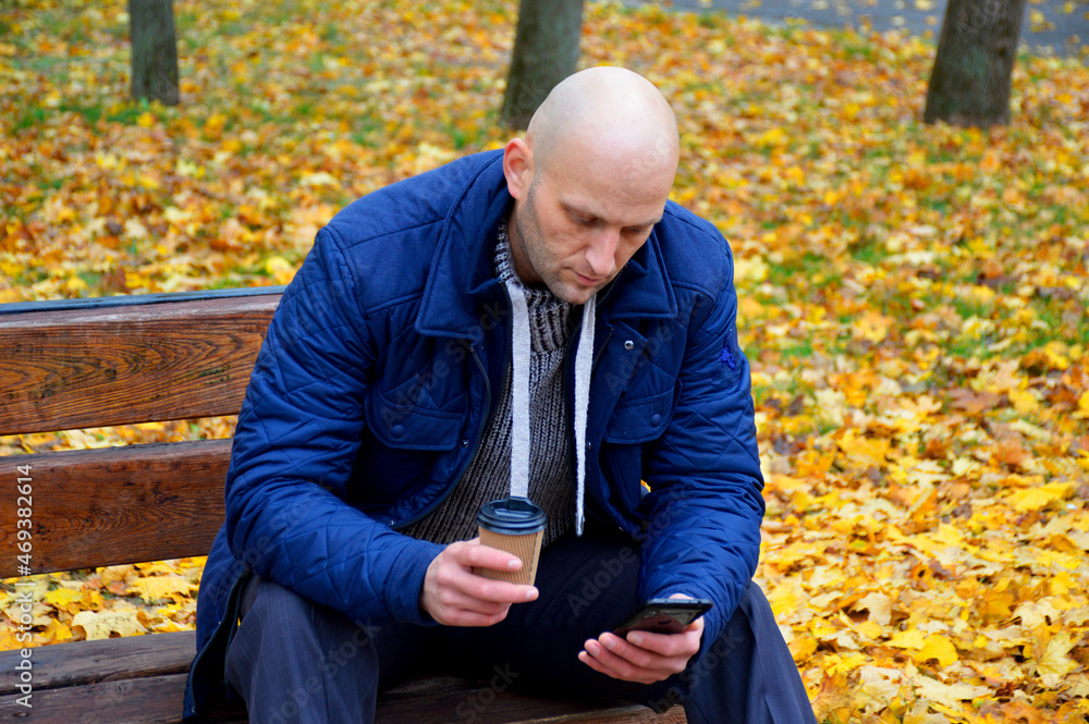 a man sits on a bench in an autumn park, holds a cup of coffee and a smartphone in his hands a man sits on a bench in an autumn park, holds a cup of coffee and a smartphone in his hands