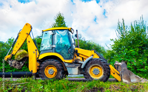 Parked large yellow multifunctional wheel tractor on the glade against green trees in sunny day. Yellow backhoe