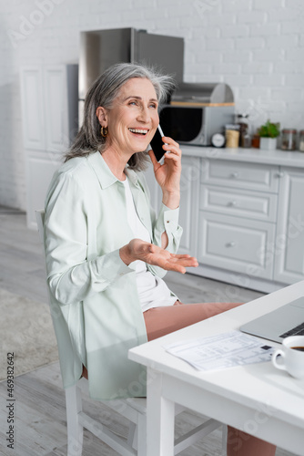 Smiling mature woman talking on smartphone near laptop  blurred coffee and bills in kitchen.