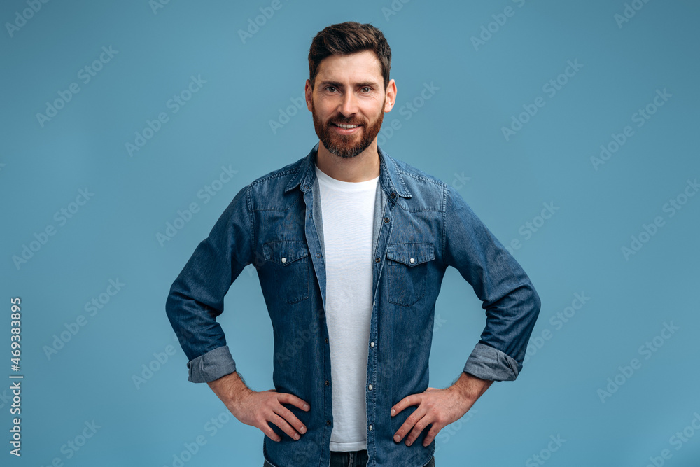 Fototapeta premium Waist up portrait view of the stylish caucasian man looking at the camera with calm confident expression. Indoor studio shot isolated on blue background