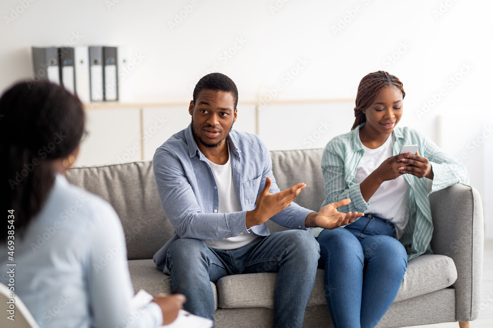 Irritated black guy complaining about his wife's smartphone addiction during psychological consultation at office Irritated black guy complaining about his wife's smartphone addiction during psychological consultation at office