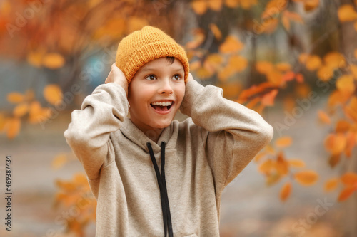  Boy with in a ginger hat in autumn day. Kid in yellow knitted hat is smilling and covering ears with hands. Behind the back of the boy magnificent yellow tree. Close up portrait.