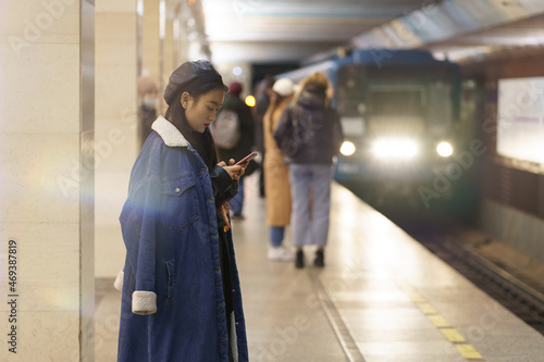 Stylish japanese girl read email in cellphone at metro station with train arriving. Young asian woman messaging with friends, posting to social media or writing comments to bloggers at subway platform photo