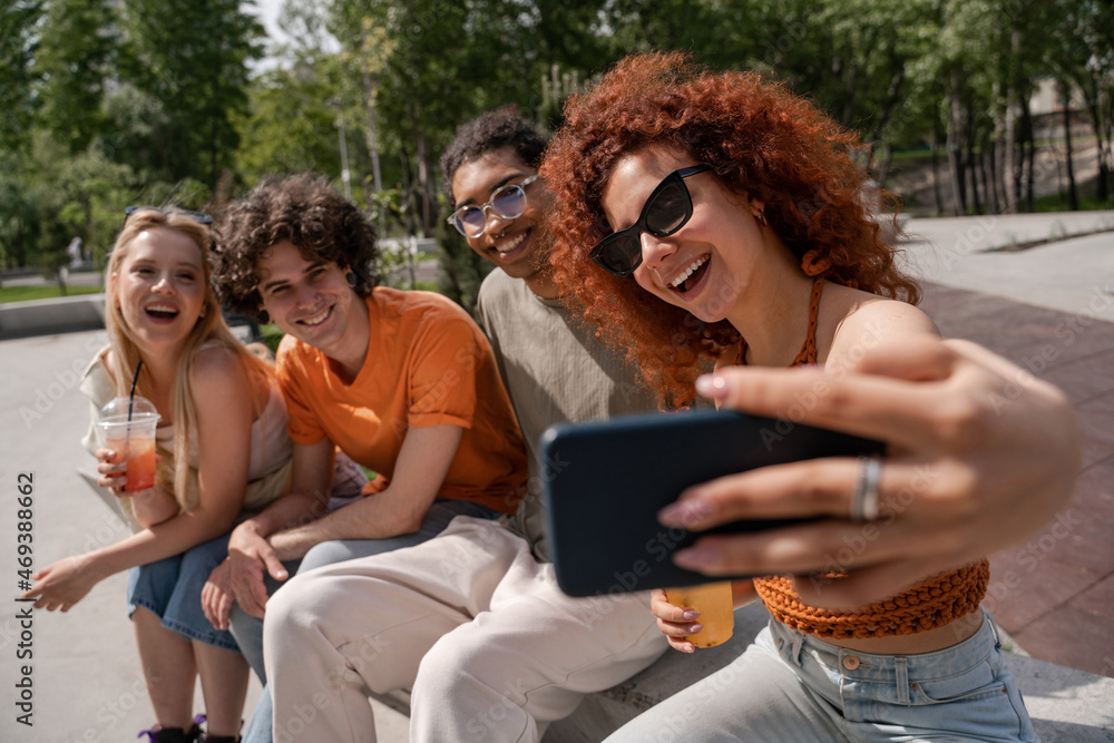 Obraz premium curly and cheerful woman in sunglasses taking selfie with multiethnic friends.