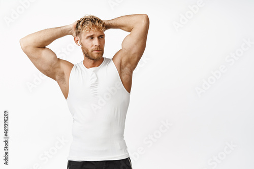 Handsome fitness guy with athletic body, holding hands behind head, flexing biceps and looking aside, workout in gym, white background