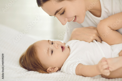 Mother and cute little child cuddling on clean white bed in nursery room or bedroom. Happy young mom playing with beautiful baby girl and looking at her with expression of love and tenderness on face