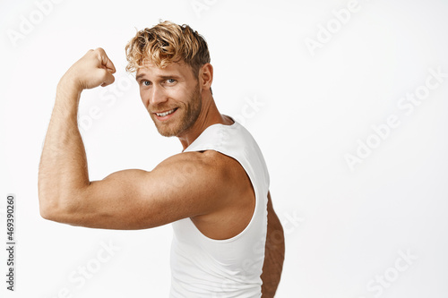 Smiling sportsman flex biceps, showing strong arm muscle, workout in gym, doing sport exercises, white background