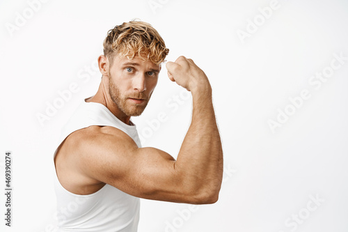 Portrait of handsome blond male athlete, fitness trainer flexing biceps, showing biceps on arm and looking like macho at camera, white background