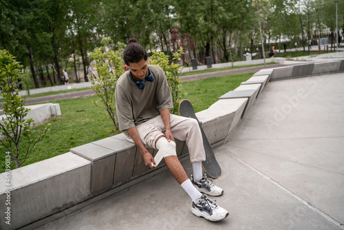 Slika na platnu young african american skater bandaging injured knee while sitting on border bench