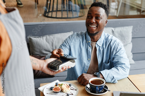 Canvas Print Joyful young Black man sitting at table in cozy cafe paying for lunch using his