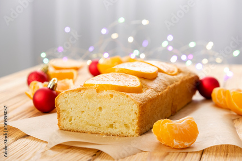 Loaf of gluten free mandarin cake with pieces of tangerine  Christmas toys and lights on rustic wooden background. Citrus pie by classic recipe for winter holidays.