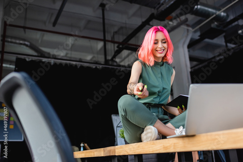 happy businesswoman with pink hair holding pen and notebook while sitting on desk and looking at laptop.