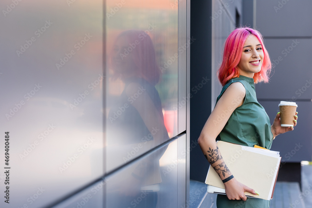 Φωτογραφία tattooed and happy businesswoman with pink hair holding folder, laptop and coffee to go outside