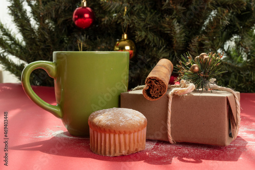 on a table a cup with hot drink, bread, gift box decorated with bow, in the background a Christmas tree with spheres, home ready to celebrate Christmas