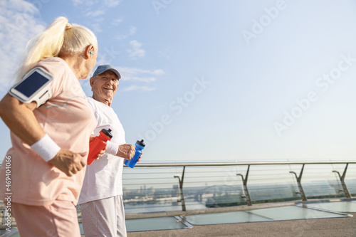 Cheerful senior man and woman with bottles of water runs along city footbridge