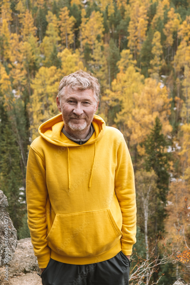 An authentic portrait of a smiling unshaven adult male traveler in a yellow hoodie on top of a mountain, against the background of an autumn forest. Selective focus. Close-up. Sustainable environment. An authentic portrait of a smiling unshaven adult male traveler in a yellow hoodie on top of a mountain, against the background of an autumn forest. Selective focus. Close-up. Sustainable environment.