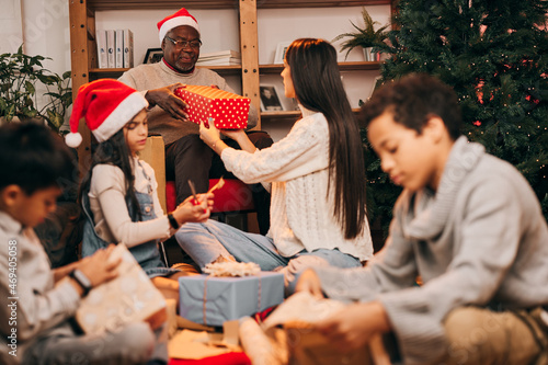 A happy woman sitting on the floor with her children and giving Christmas gift to her father. Multiethnic family celebrating Christmas at home.