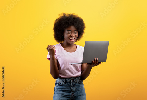 Euphoric african american lady celebrating success and holding laptop, raising clenched fist over yellow background