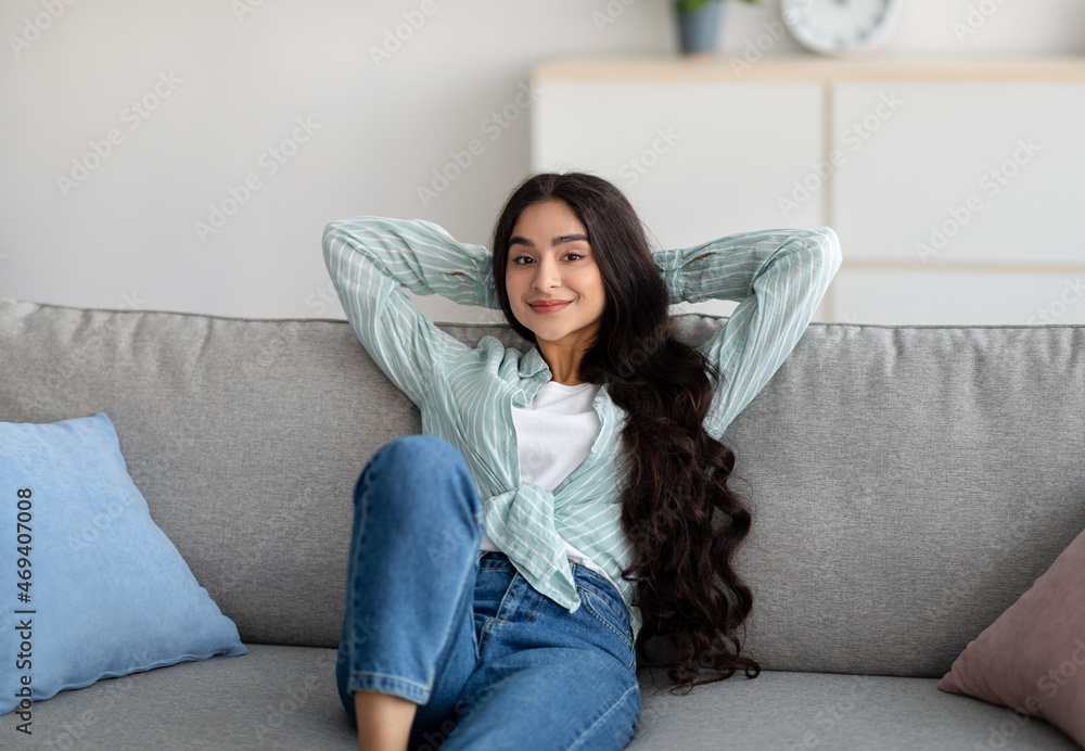 Young beautiful Indian lady relaxing on weekend morning at home, sitting on sofa, holding hands behind head Young beautiful Indian lady relaxing on weekend morning at home, sitting on sofa, holding hands behind head