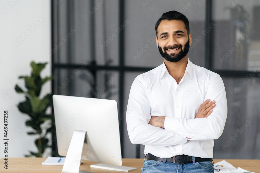 Portrait of a successful intelligent Indian businessman, manager or IT specialist, wearing a white shirt, standing near his desk in the office with arms crossed, looking at the camera with a friendly - obrazy, fototapety, plakaty 